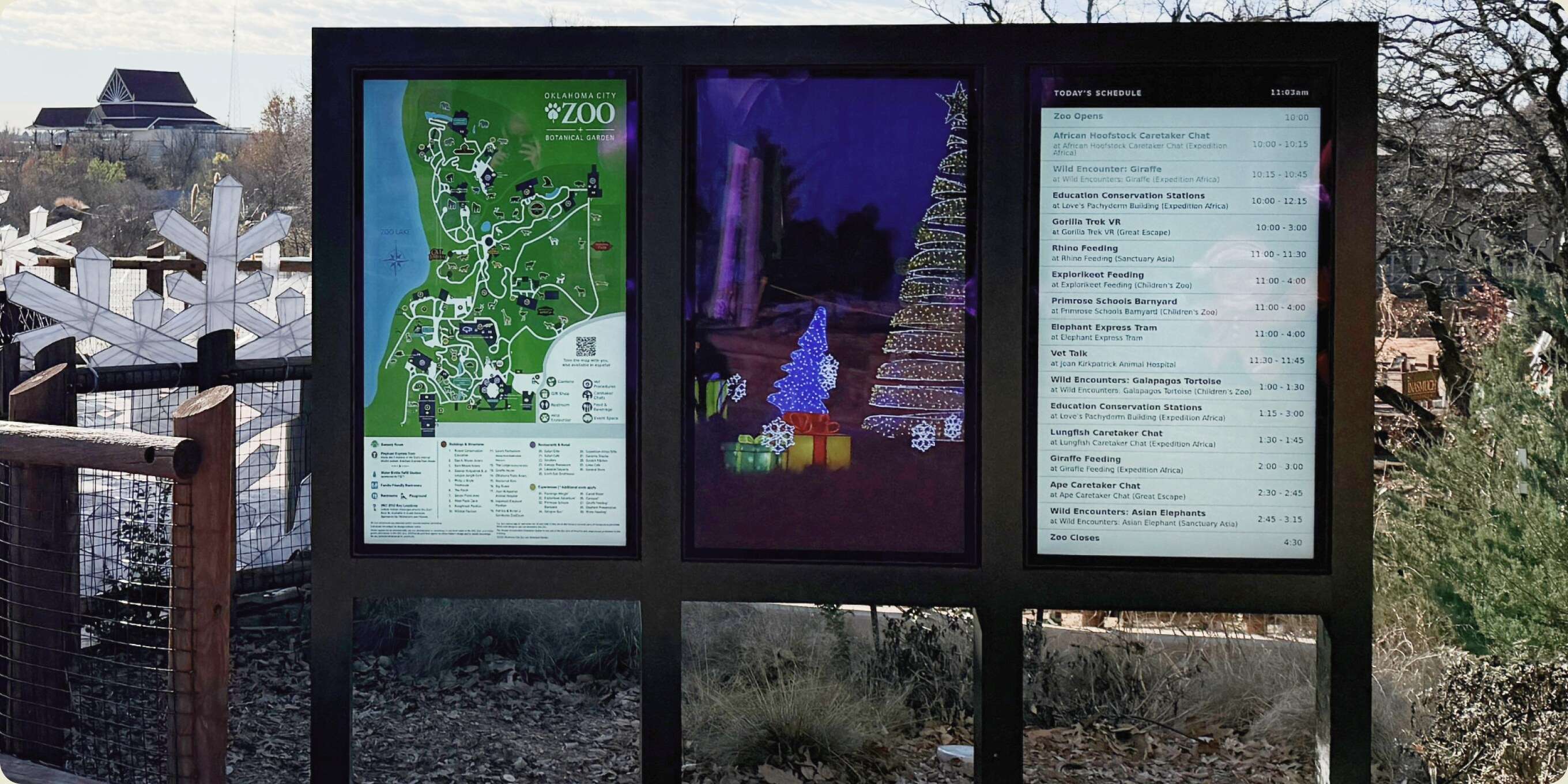 An outdoor kiosk at the zoo carrying three digital signs — the park map, a seasonal lights promotion, and a schedule — beside a shaded shelter on a winter afternoon.